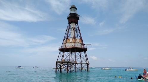 A weathered lighthouse stands on stilts in turquoise waters, with boats and swimmers nearby under a blue sky.