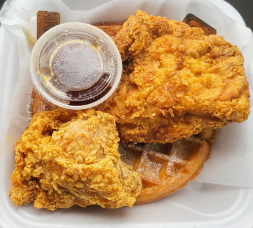 A takeout container with two pieces of fried chicken on a waffle, accompanied by a small cup of syrup.