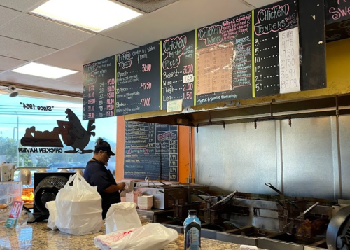 A restaurant counter with a menu board, a cook preparing food, and takeout bags on the counter.