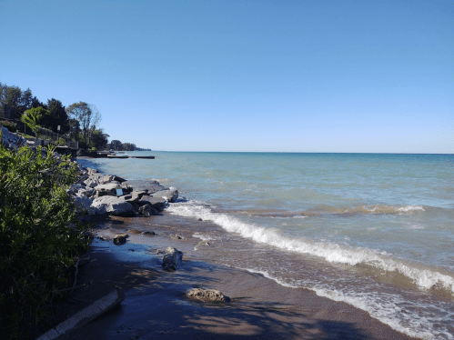 A serene lakeshore scene with gentle waves, rocky shoreline, and clear blue skies.