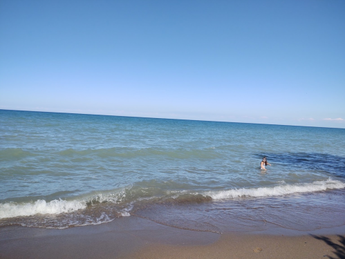 A person wades in the calm, blue waters of a beach under a clear sky. Gentle waves lap at the sandy shore.