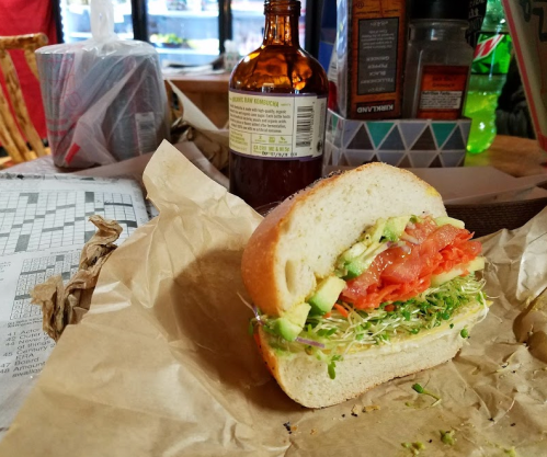 A sandwich with sprouts, tomato, and avocado on a paper wrapper, with a bottle and cluttered table in the background.