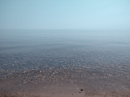 Calm, clear water meets a sandy shore under a hazy sky, creating a serene coastal scene.