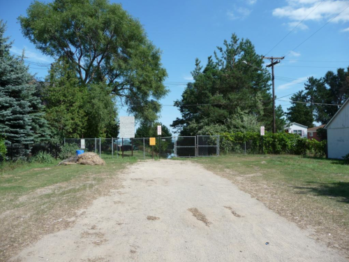 A dirt path leads to a fenced area surrounded by trees and greenery under a clear blue sky.