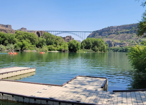 A serene lake with green trees, wooden docks, and a bridge in the background under a clear blue sky.