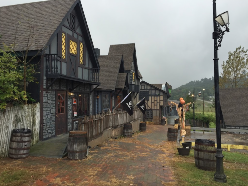 A quaint street scene featuring dark, Tudor-style buildings, barrels, and a scarecrow under a cloudy sky.