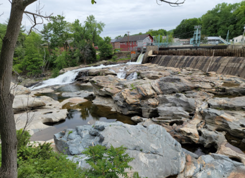 A rocky river landscape with a small waterfall, surrounded by greenery and a building in the background.
