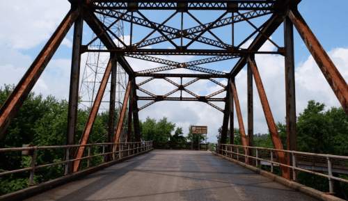 Rusty metal bridge with a lattice design, surrounded by green trees and a blue sky with clouds.