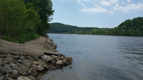 A serene river scene with rocky banks, lush greenery, and gentle hills under a partly cloudy sky.