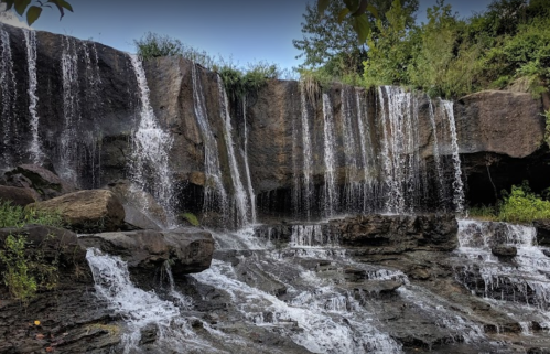 A serene waterfall cascading over rocky ledges, surrounded by lush greenery and a clear blue sky.