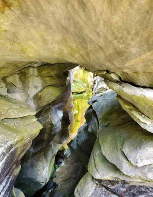 A narrow rock passageway with moss-covered walls, leading to a green, illuminated area beyond.