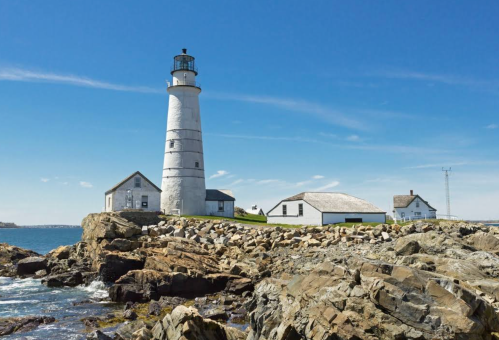 A tall white lighthouse stands on rocky shores, surrounded by small buildings under a clear blue sky.
