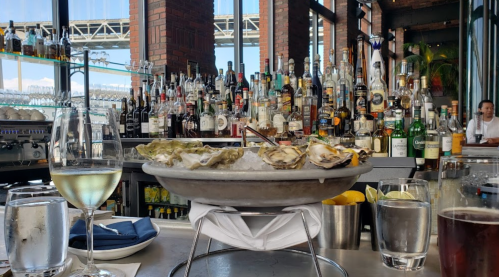 A seafood platter with oysters on ice, surrounded by drinks and a bar filled with various bottles in a restaurant setting.