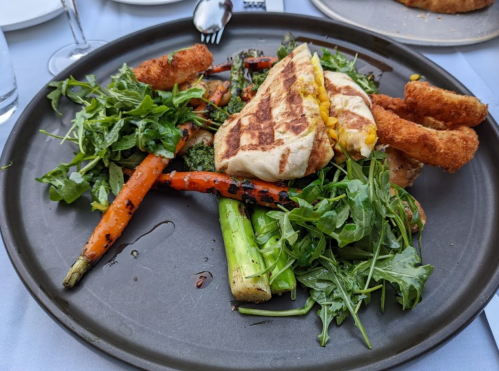 A plate of grilled vegetables, including carrots and greens, topped with a sandwich and crispy onion rings.