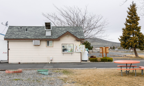 A small, single-story building with a drive-thru window, surrounded by gravel and grass, under a cloudy sky.