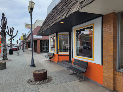 A street view of a small town with benches, shops, and a tree-lined sidewalk under a cloudy sky.