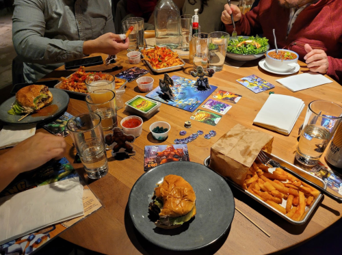 A table filled with various dishes, drinks, and board game pieces, with people enjoying a meal together.