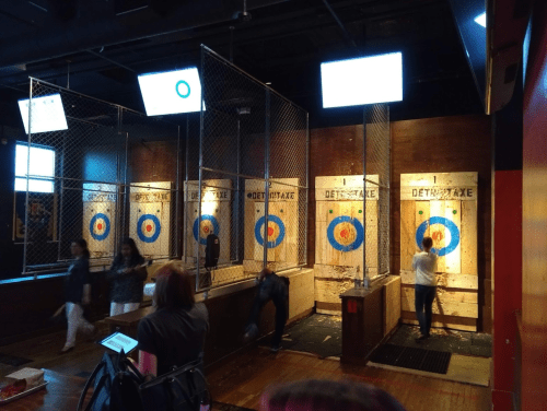 Axe-throwing venue with multiple targets, people throwing axes, and screens displaying scores.
