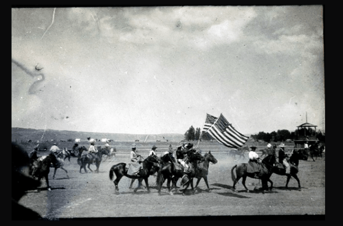 Black and white photo of a parade with riders on horseback carrying an American flag. Dust clouds in the background.
