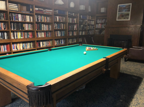A green felt pool table in a cozy room with bookshelves and a fireplace in the background.