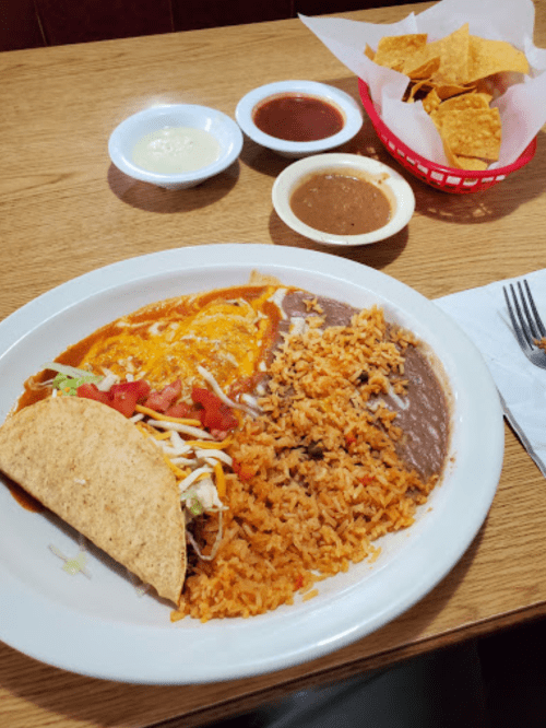 A plate of enchiladas, taco, rice, and refried beans, with a side of chips and sauces in the background.