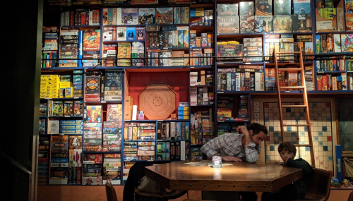A man and a child sit at a table in front of a wall filled with board games. A ladder leans against the shelf.