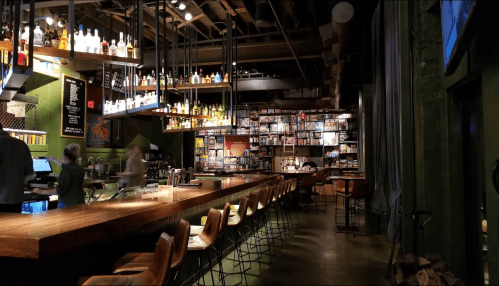 A cozy bar with a wooden counter, shelves of liquor, and a wall of board games, featuring a few patrons and staff.