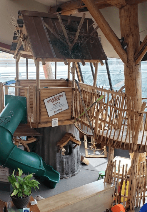 A wooden play structure resembling a treehouse with a slide, bridge, and natural decor in a bright indoor space.