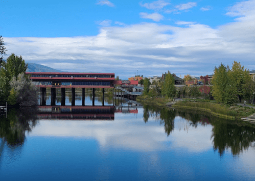 A serene river scene with a modern building on stilts, surrounded by trees and reflecting clouds in the water.
