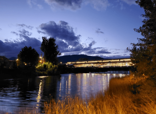 A serene river scene at dusk, with reflections of lights and clouds in the water, surrounded by trees and grass.