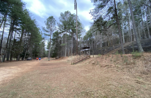 A grassy area near a sports field surrounded by trees, with a set of bleachers and a few people walking in the distance.