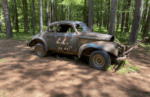 An old, rusty car sits abandoned in a forest, surrounded by trees and dappled sunlight on the ground.