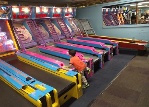 A young child plays at a colorful arcade, focused on a skee-ball game with multiple lanes in the background.