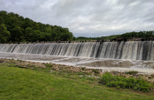 A wide waterfall cascading over a dam, surrounded by green trees and a cloudy sky.