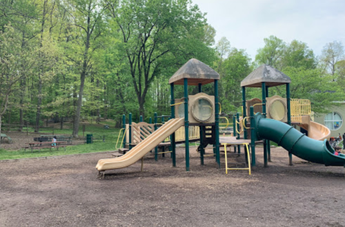 A playground with slides and climbing structures surrounded by trees and a dirt area.