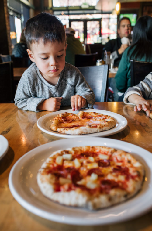 A young boy sits at a table, focused on a pizza in front of him, with another pizza nearby in a busy restaurant.