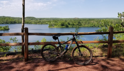 A mountain bike rests against a wooden fence overlooking a serene lake surrounded by lush greenery.