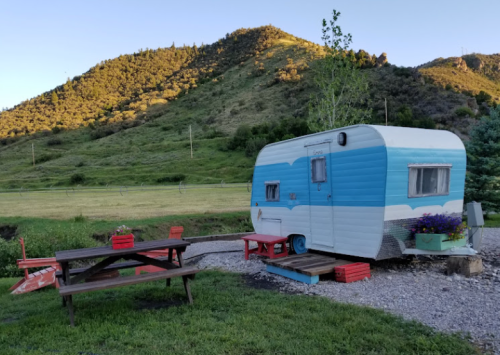 A vintage blue and white camper trailer beside a picnic table, set against a green hillside under a clear sky.