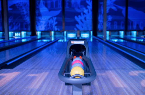 A bowling alley with colorful balls in a rack, illuminated by blue lighting and decorated with ocean-themed murals.