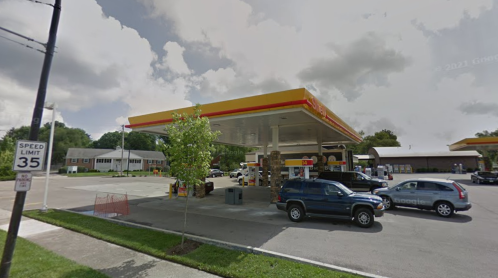 A Shell gas station with several cars parked, surrounded by trees and a residential area under a cloudy sky.