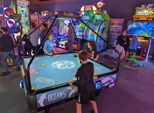 Two children play air hockey at an arcade, surrounded by colorful games and other players in the background.