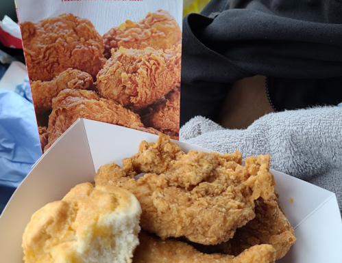 A close-up of fried chicken pieces and a biscuit in a takeout box, with a menu featuring fried chicken in the background.