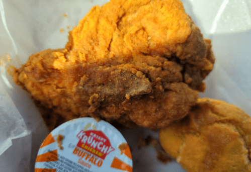 A piece of crispy fried chicken with a side of buffalo sauce and a biscuit in a takeout container.