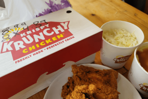 A box of Krispy Krunchy Chicken with fried chicken pieces, coleslaw, and a cup of gravy on a wooden table.