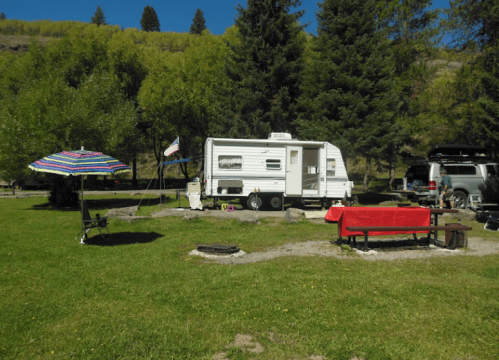 A campsite with a white RV, picnic table, umbrella, and fire pit surrounded by trees and grass.