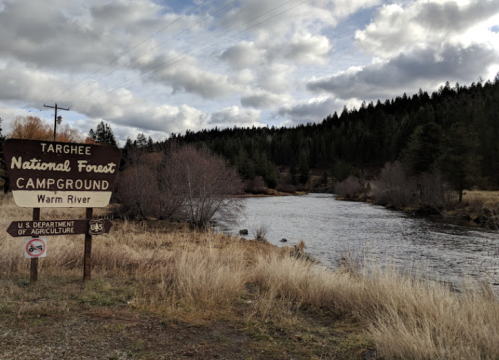 Sign for Targhee National Forest Campground by the Warm River, surrounded by trees and grassy areas under a cloudy sky.