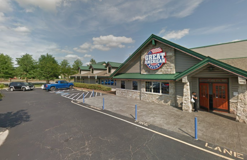 Exterior view of Great American Cookies building with parking lot and blue truck, surrounded by trees and a clear sky.