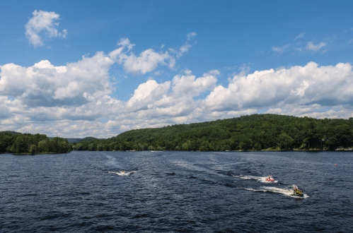 Three jet skis glide across a calm lake under a blue sky with fluffy clouds, surrounded by green hills.