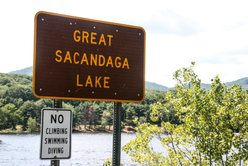 Sign for Great Sacandaga Lake with a warning sign below prohibiting climbing, swimming, and diving. Trees and water in background.