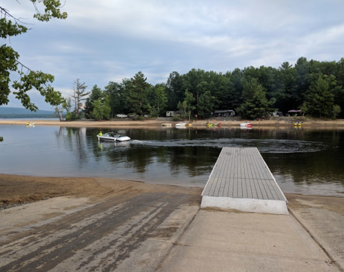 A calm lake scene with a boat approaching a dock, surrounded by trees and a sandy beach in the background.